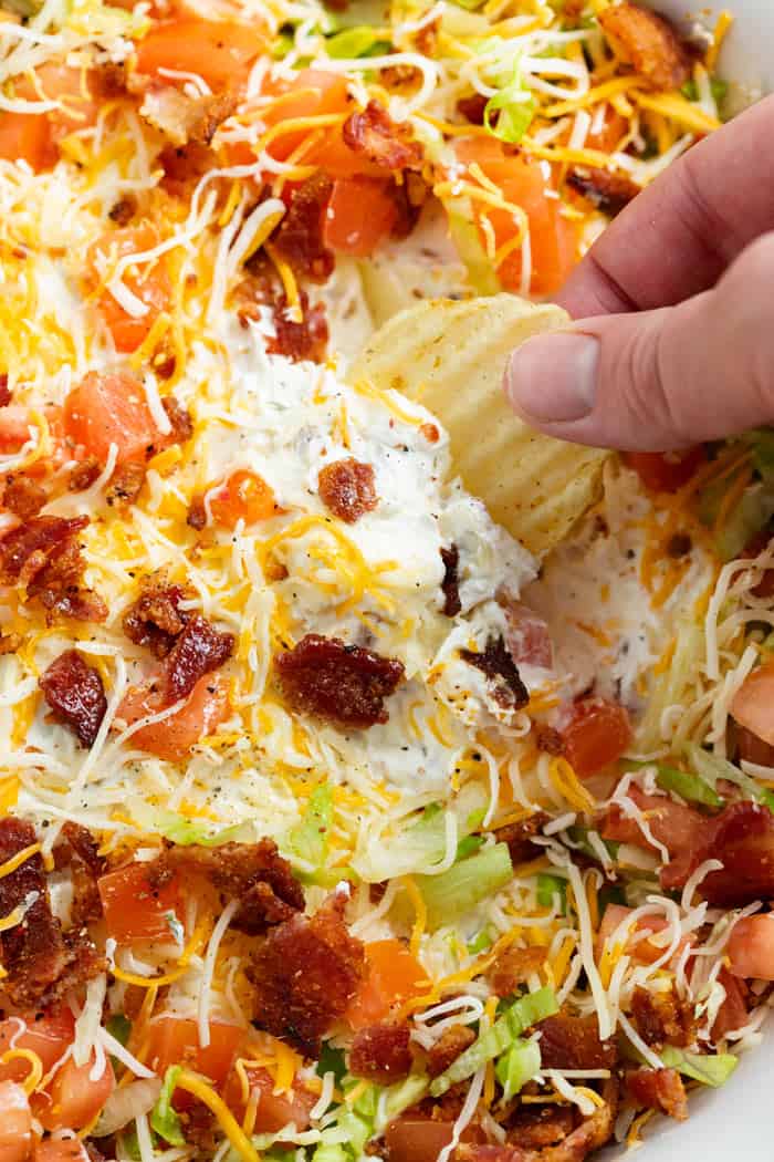 A hand dipping a potato chip into BLT Dip with Bacon, Lettuce, and Tomatoes on top.