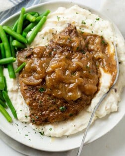 A pile of mashed potatoes topped with cubed steak with brown gravy and onions with green beans on the side.