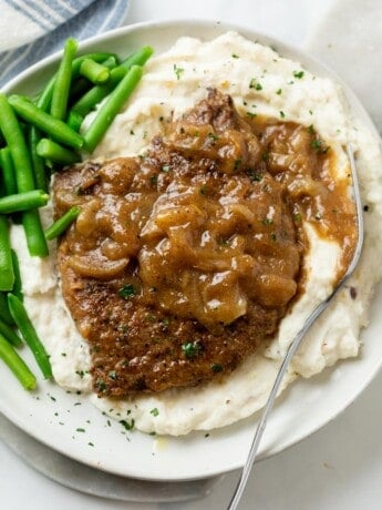 A pile of mashed potatoes topped with cubed steak with brown gravy and onions with green beans on the side.