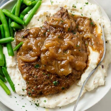A pile of mashed potatoes topped with cubed steak with brown gravy and onions with green beans on the side.