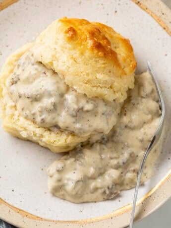 A buttermilk biscuit slice in half and topped with Sausage Gravy with a fork on the side.