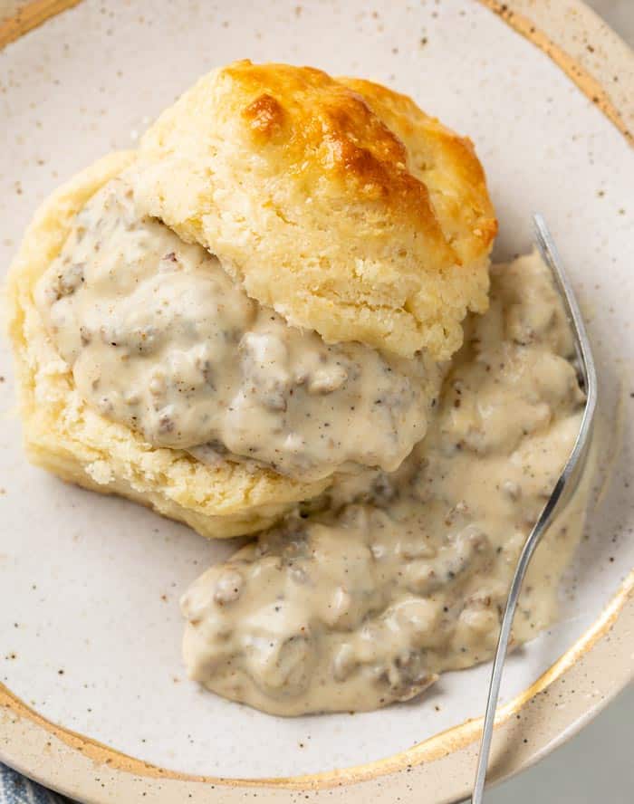 A buttermilk biscuit slice in half and topped with Sausage Gravy with a fork on the side.
