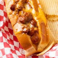 A chili cheese dog on a red and white checkered paper with potato chips in the background.