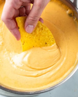 A hand dipping a tortilla chip into nacho cheese sauce.