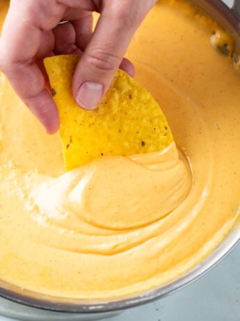 A hand dipping a tortilla chip into nacho cheese sauce.