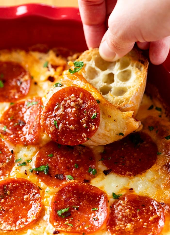 A hand dipping a slice of French bread into Pizza Dip with Pepperoni.