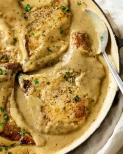 A skillet with Pork Chops in Gravy with a spoon on the side and fresh parsley on top.