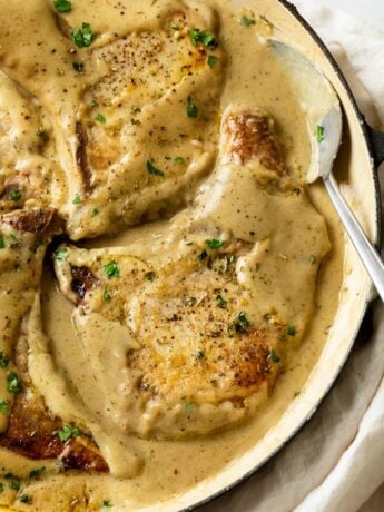 A skillet with Pork Chops in Gravy with a spoon on the side and fresh parsley on top.