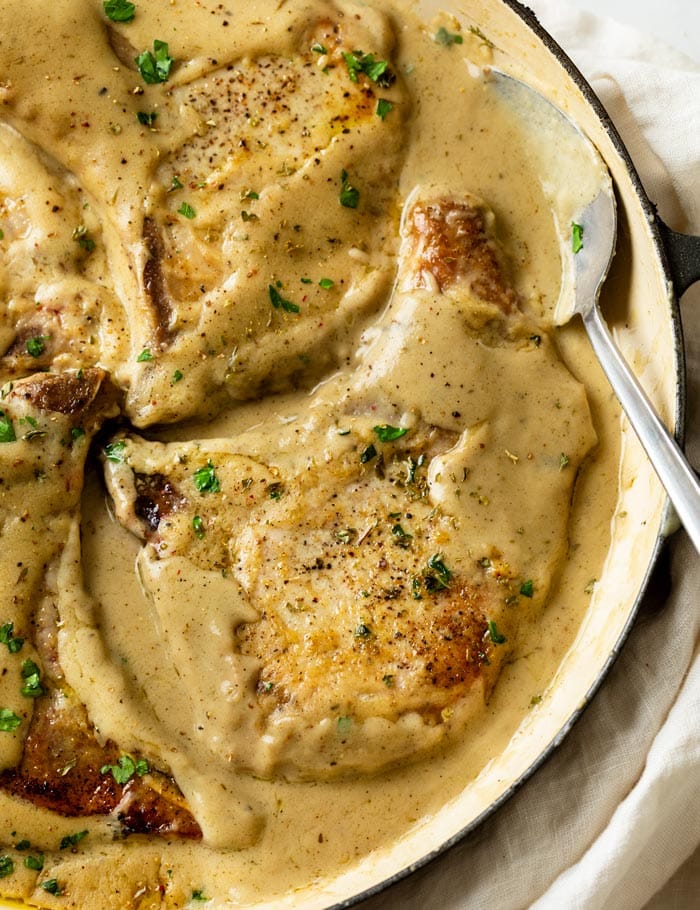 A skillet with Pork Chops in Gravy with a spoon on the side and fresh parsley on top.