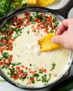 A hand dipping a tortilla chip into a skillet of White Queso Dip topped with Pico De Gallo and Cilantro