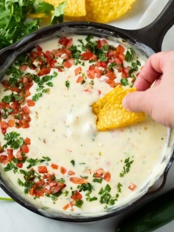 A hand dipping a tortilla chip into a skillet of White Queso Dip topped with Pico De Gallo and Cilantro
