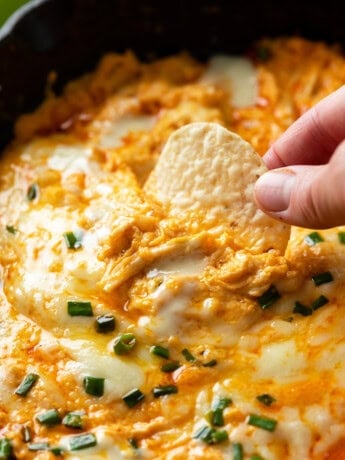 A hand dipping a Tortilla Chip into Buffalo Chicken Dip topped with cheese and chives.