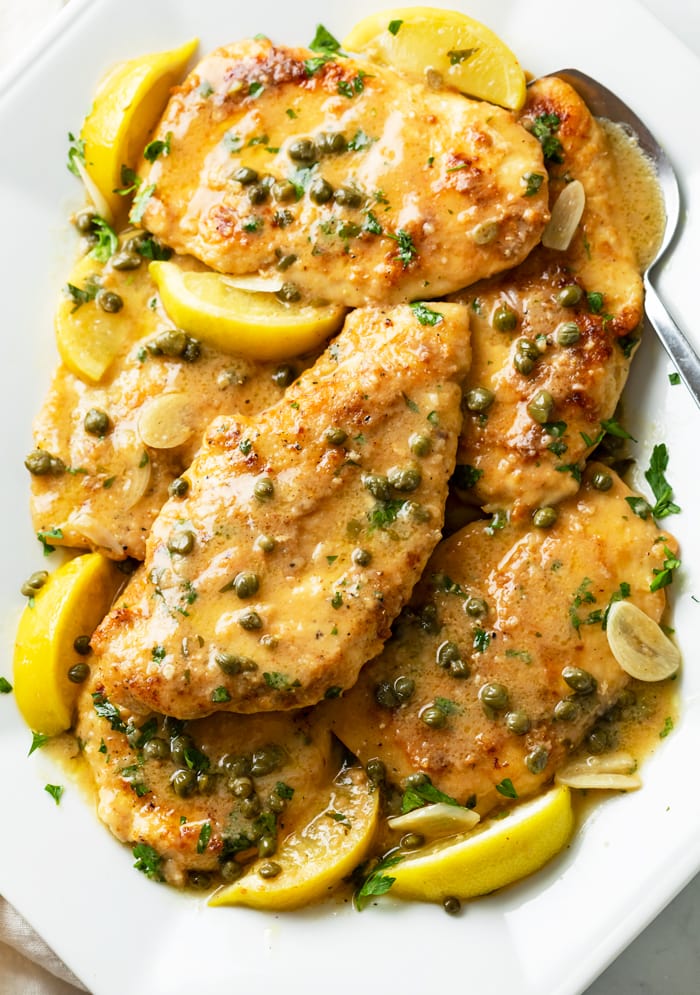 A white platter with Chicken Piccata and lemon slices with lemon sauce and capers.