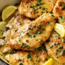 Chicken Piccata in a pot with a lemon butter sauce topped with capers, lemon wedges, and fresh parsley.