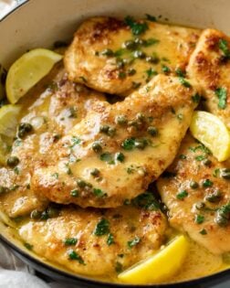 Chicken Piccata in a pot with a lemon butter sauce topped with capers, lemon wedges, and fresh parsley.