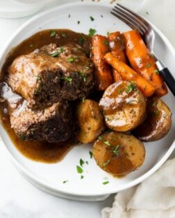 A white plate with Crock Pot Roast with gravy on top with potatoes and carrots.