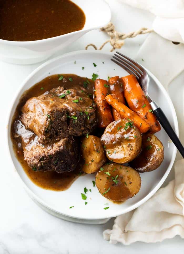 A white plate with Crock Pot Roast with gravy on top with potatoes and carrots.