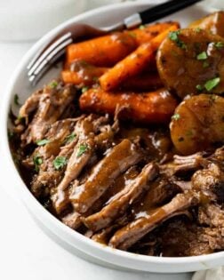 A white plate with tender Crock Pot Roast with gravy and carrots and potatoes in the background.