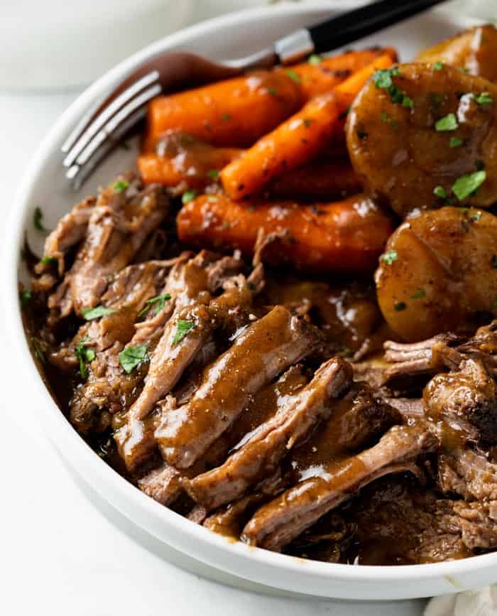 A white plate with tender Crock Pot Roast with gravy and carrots and potatoes in the background.