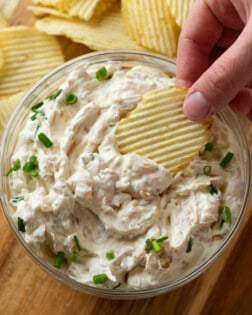 A hand dipping a Ruffled chip into a glass bowl of French Onion Dip topped with chives.