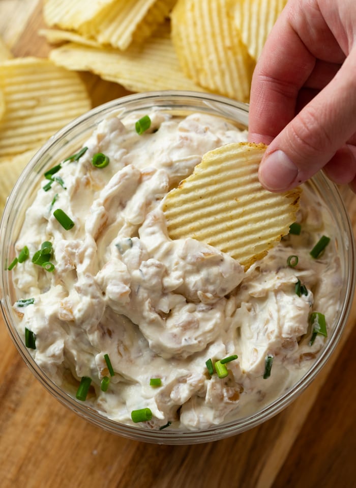 A hand dipping a Ruffled chip into a glass bowl of French Onion Dip topped with chives.