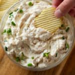 A potato chip scooping up French onion dip from a glass bowl with green onions on top.