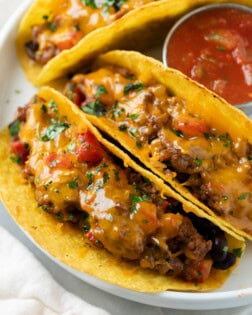 A white plate with baked tacos filled with ground beef and cheese with tomatoes.