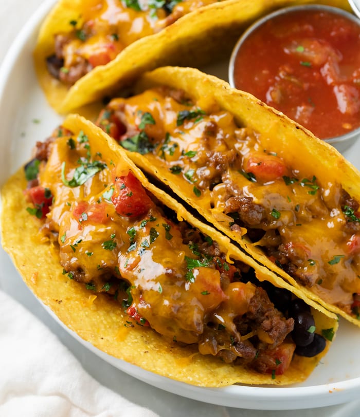 A white plate with baked tacos filled with ground beef and cheese with tomatoes.