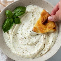 A bowl full of Feta Dip with a hand dipping bread into it with a garnish of basil.