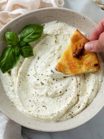 A bowl full of Feta Dip with a hand dipping bread into it with a garnish of basil.