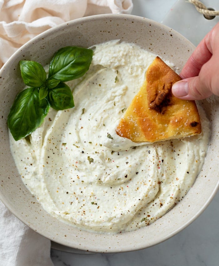 A bowl full of Feta Dip with a hand dipping bread into it with a garnish of basil.