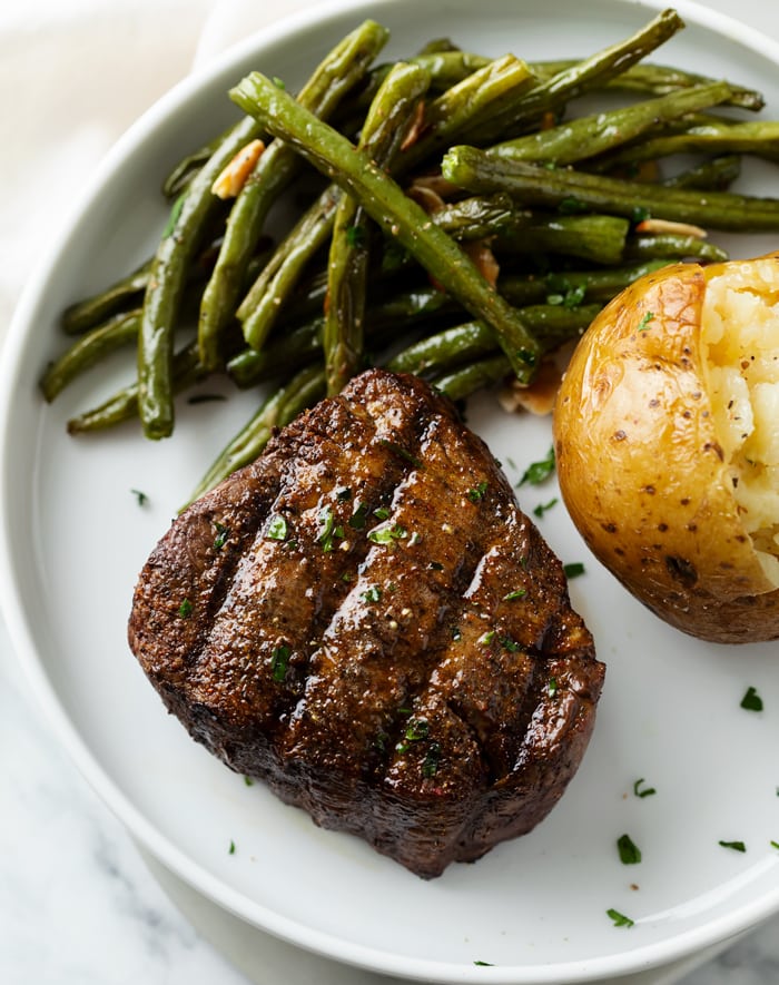 A filet mignon with steak rub on top next to roasted green beans and a baked potato.