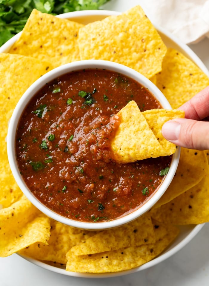 A hand dipping a tortilla chip into a white bowl of salsa with tortilla chips around it.
