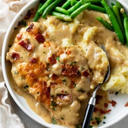 A white plate with Smothered Chicken next to mashed potatoes with gravy and green beans.
