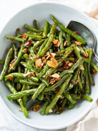 A white bowl with roasted green beans and almonds with a spoon on the side.