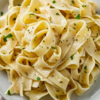 A pile of buttered noodles on a plate with herbs and Parmesan Cheese.