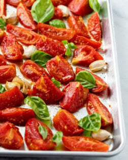 A rimmed baking sheet with roasted tomatoes and basil on top with salt and freshly cracked pepper.