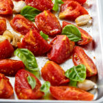Roasted tomatoes on a baking sheet with fresh basil and garlic cloves.