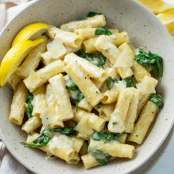 A white bowl filled with Creamy Spinach Pasta with ziti noodles and lemon wedges on the side.