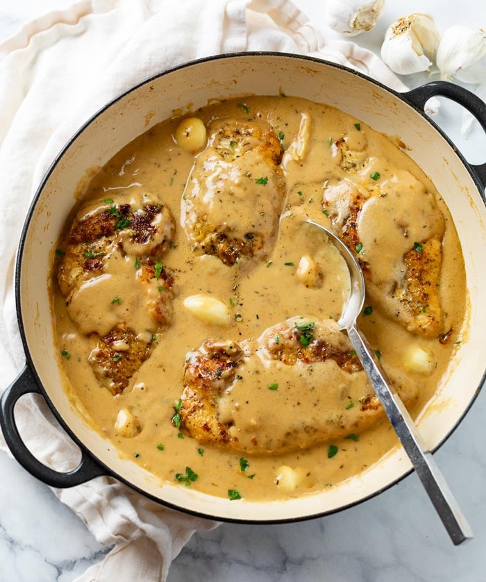 A skillet full of creamy garlic chicken with whole garlic cloves and a spoon.