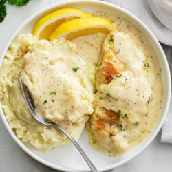 Creamy Herb Chicken on a white plate with mashed potatoes, cream sauce, and lemon wedges.