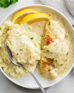 Creamy Herb Chicken on a white plate with mashed potatoes, cream sauce, and lemon wedges.