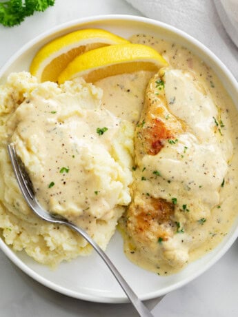 Creamy Herb Chicken on a white plate with mashed potatoes, cream sauce, and lemon wedges.