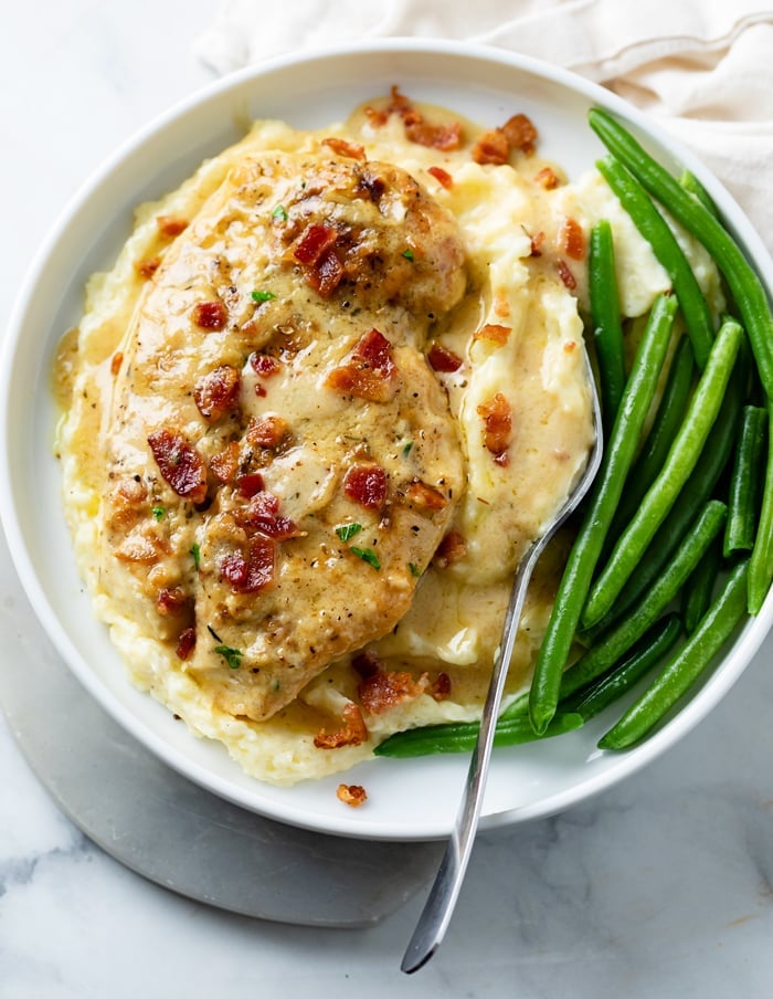 A white plate with Creamy Bacon Chicken on top of mashed potatoes with green beans.