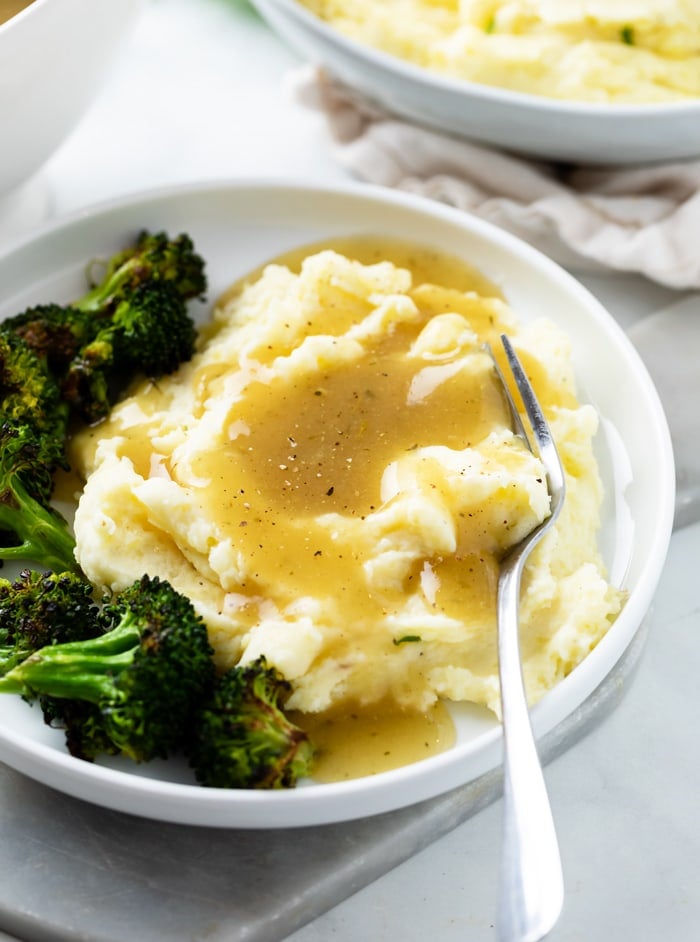 A white plate with a pile of mashed potatoes with chicken gravy on top and broccoli on the side.