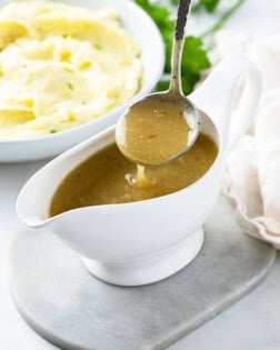 A ladle scooping up chicken gravy from a gravy boat with mashed potatoes in the background.