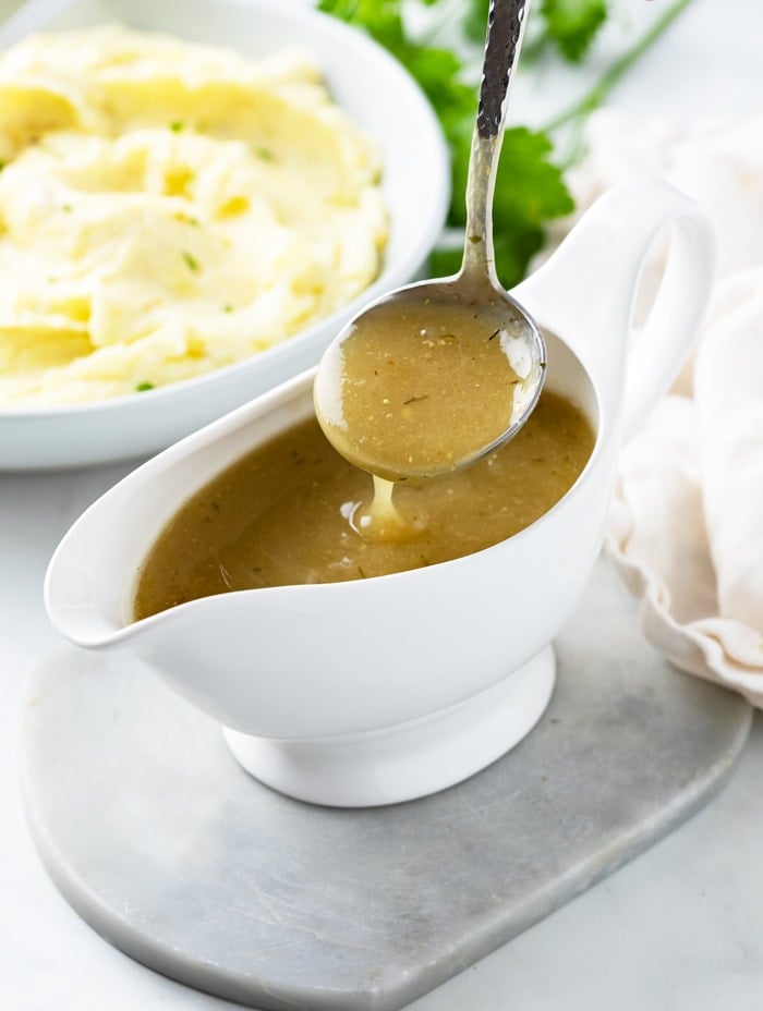 A ladle scooping up chicken gravy from a gravy boat with mashed potatoes in the background.