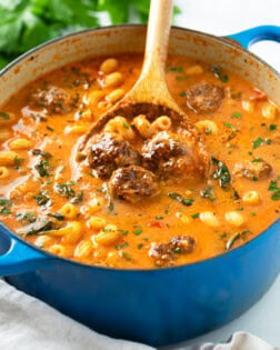 A blue dutch oven filled with Meatball Soup with meatballs, pasta, spinach, and parsley.