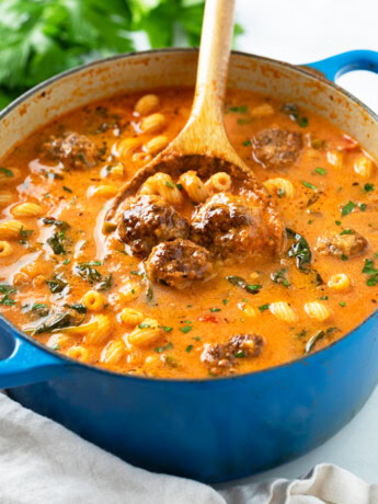 A blue dutch oven filled with Meatball Soup with meatballs, pasta, spinach, and parsley.