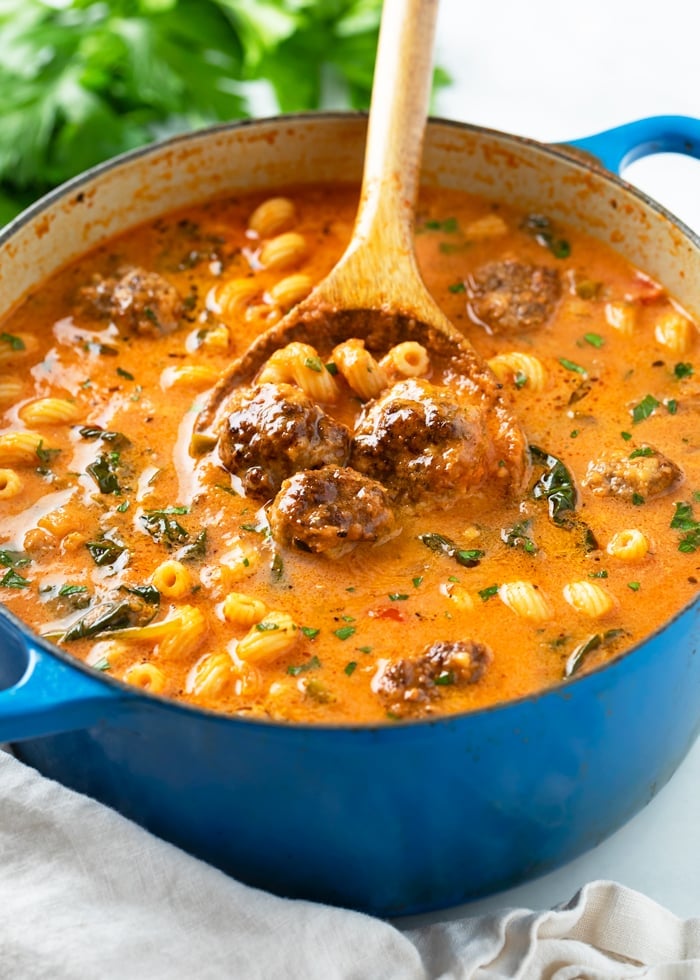 A blue dutch oven filled with Meatball Soup with meatballs, pasta, spinach, and parsley.